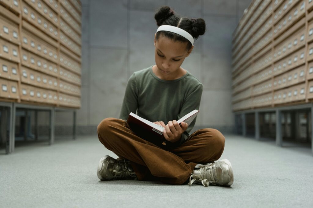 A young girl sitting cross-legged, reading a book in a library with card catalogs.